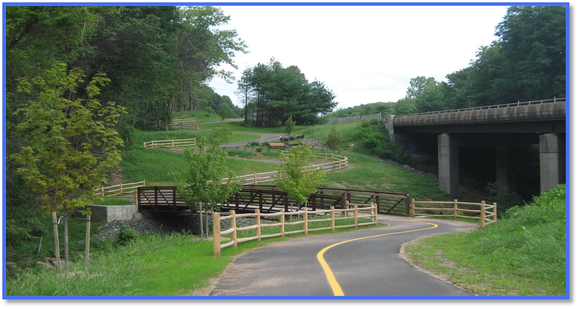 "A paved multi‑use trail curves through a grassy landscape with wooden railings on both sides. The trail crosses a small wooden bridge over a stream. Additional wooden fences line the hillside in the background. A large highway bridge stands to the right of the trail, supported by concrete columns. Trees and greenery surround the area."